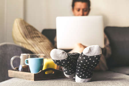 Woman sitting on a couch in the living room with warm socks in a winter morning. Girl using laptop and works at home, having natural breakfast with tea coffee and fruits. Focus on feet in the foregroundの写真素材