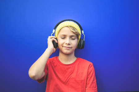 Portrait of a smiling beautiful boy listening music with headphones isolated on a blue background. Colorful image of young male wearing a wool cup, have fun with earphones. Youth fun isolation conceptの写真素材