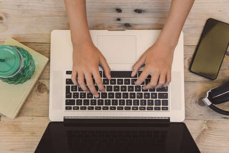 Hands of a young boy typing on a laptop keyboard over a rustic wooden table. Teen using a computer to download good music on his smartphone. Teenager sharing multimedia content with friends on line.の写真素材