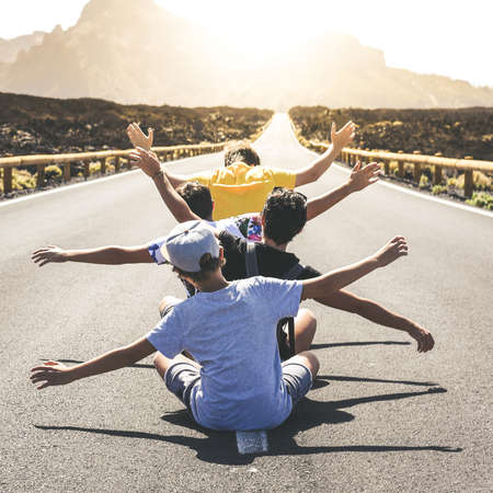 Group of teenager sitting in the middle of a long road exulting with raising arms. Back view of four teen enjoying free time on holiday with open arms. Youth, freedom, travel and adventure concept.の写真素材