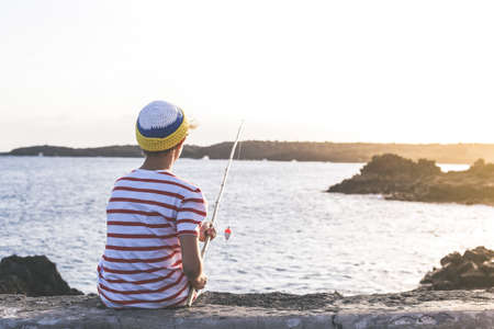 Back view of a young fisherman with with fishing pole in hand looking the sea. Sailor boy with wool cap and a striped t-shirt enjoying a summer evening. Lifestyle, outdoors, fun and carefree conceptの写真素材