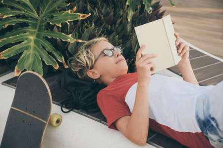 Young boy reading sitting outside in a beautiful tropical garden. Happy student doing homework at the park Child with book in hand relaxing outdoors Educational youth modern alternative school conceptの写真素材