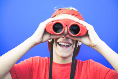 Beautiful boy looking away with a red binoculars isolated on a blue background. Colorful image of a young male having fun with a telescope. Fun, astonishment, happiness concept. Focus on the mouth.の写真素材