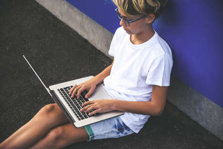 Portrait of a trendy boy using and laptop sitting outdoors. Teen doing homework online outside...