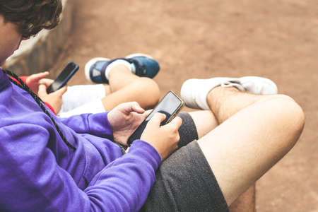 Close up view of a couple of teen using smartphone sitting outdoor at the park. Teenagers...