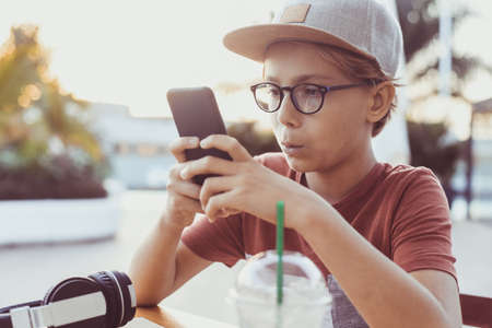Teenager watching video on smartphone sitting at the table. Young boy drinks smoothie outside at the bar using cell phone. Teen using mobile phone reading friends chat. New tech and youth conceptの写真素材