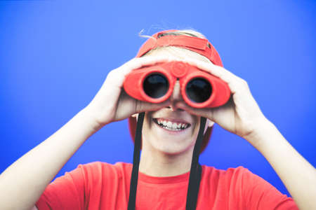 Beautiful boy looking away with a red binoculars isolated on a blue background. Colorful image of a young male having fun with a telescope. Fun, astonishment, happiness concept. Focus on the mouth.の写真素材