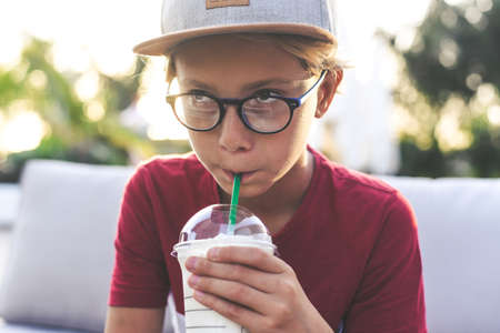 Trendy young boy drinking a smoothie sitting outdoor at the bar. Teen with hat drinks a cold beverage outside in the garden. Portrait of pensive teenager enjoy a summer day Happiness free time conceptの写真素材