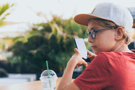 Teenager watching video on smartphone sitting at the table. Young boy drinks smoothie outside at the...