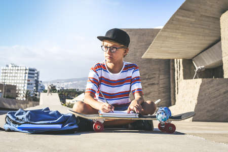 Trendy student doing homework out of the college sitting on a concrete wall. Young boy writing on an exercise book using a skateboard like a desk in a urban context. Educational school, youth conceptの写真素材