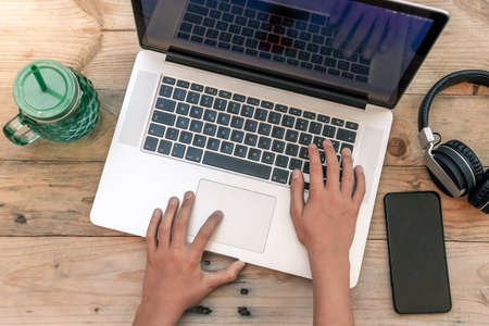 Hands of a young boy typing on a laptop keyboard over a rustic wooden table. Teen using a computer to download good music on his smartphone. Teenager sharing multimedia content with friends on line.の写真素材