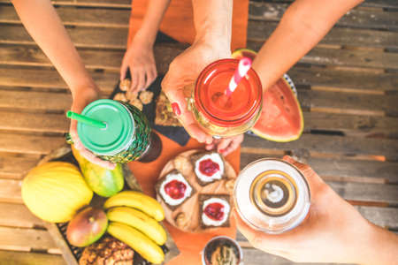 Hands toasting with natural drinks in colorful glasses with straws. Wooden table with natural food, in the background for a healthy and nutritious breakfast. Vegan, vegetarian, bio, diet, meal conceptの写真素材