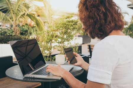 Woman working with laptop and smartphone sitting outdoor in a beautiful terrace. Female using computer and cellphone sitting in a bar dehor drinking coffee. Home work, business and technology conceptの写真素材