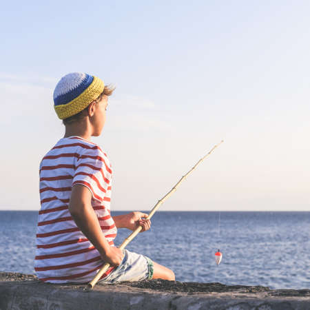 Back view of a young fisherman with with fishing pole in hand looking the sea. Sailor boy with wool cap and a striped t-shirt enjoying a summer evening. Lifestyle, outdoors, fun and carefree conceptの写真素材