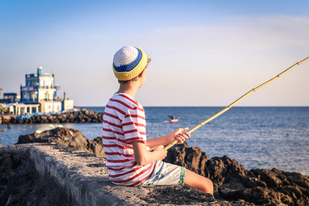 Back view of a young fisherman with with fishing pole in hand looking the sea. Sailor boy with wool cap and a striped t-shirt enjoying a summer evening. Lifestyle, outdoors, fun and carefree conceptの写真素材