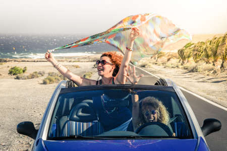 Summer vacation and travel concept image of a young woman waving headscarf out of hood of convertible car Curly brunette girl arms up having fun in a cabriolet topless. Female friends enjoying freedomの写真素材