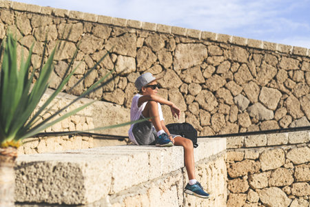 Smiling trendy boy enjoying sunny day outdoor. Portrait of a caucasian teen sitting on a stone wall. Cool teenager casual dressed with rapper hat, looking away. Youth, carefree, happiness concept.の写真素材