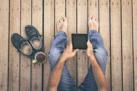 Top view of a man reading an e-book on digital tablet device barefoot sitting on a wooden floor. Cup of coffee and fashion sneakers on the side. Boy enjoying new technology. Leisure tech trend conceptの写真素材