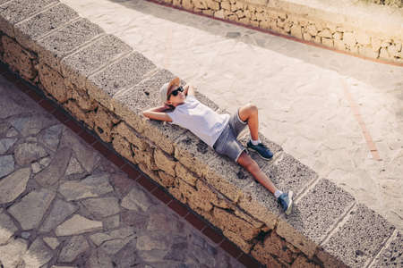 Young trendy boy enjoying sunny day outdoor lying on a stone wall in the city .. Portrait of a...