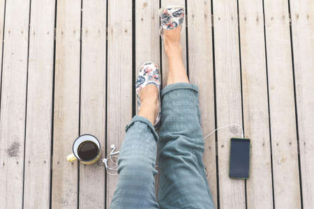 Top view of female legs with trendy trousers and floral shoes. Cup of coffee and smartphone with...