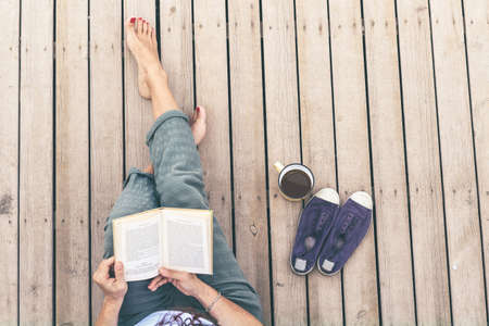 Top view of a woman reading a book, barefoot, sitting on a wooden floor. Cup of coffee and fashion sneakers on the side. Girl dressed casual enjoying free time. Female, leisure and relax concept.の写真素材