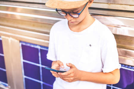 Beautiful trendy boy using smartphone in a urban context. Image of kid playing with mobile phone. Caucasian blonde young man communicate with friends. Youth, new trend technology, online concept.の写真素材