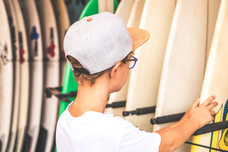 Beautiful trendy boy looking for a surf to enjoying ocean waves with friends. Young surfer choose the new board for the school training. Sporty young man ready to go to the beach. Youth, sport conceptの写真素材