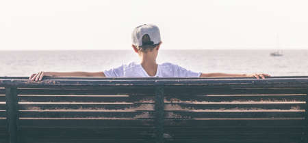 Boy alone sitting on a bench in front of the sea watching the horizon. Sad young tourist waiting the...