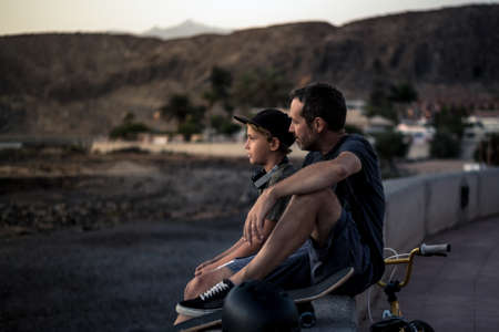 A teen and his father sitting on a wall with bicycle and skateboard. Young rider and dad relaxing after a fun day of jump and tricks with bm. People enjoying a summer evening, exotic backgroundの写真素材