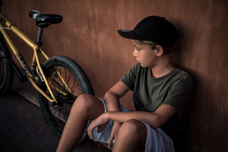 Beautiful trendy boy relaxing alone sitting near his bicycle. Young man looking a bmx after riding on a summer day before start of the school. Portrait of a kid with rapper hat. Youth fun concept.の写真素材