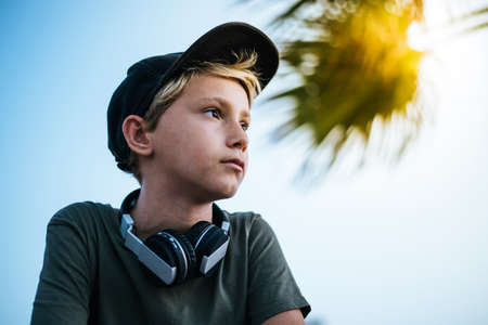 Portrait of a happy trendy boy thinking serious. Pensive blonde young boy with hat and headphones. Warm filter, exotic background. Teenager enjoying outdoor, Youth, recreation, free time conceptの写真素材
