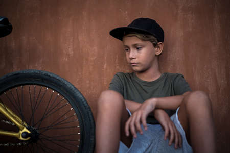 Beautiful trendy boy relaxing alone sitting near his bicycle. Young man looking a bmx after riding on a summer day before start of the school. Portrait of a kid with rapper hat. Youth fun concept.の写真素材