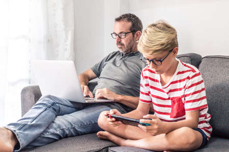 Young boy playing with a video game console while his dad is working with laptop, sitting on a sofa at home. Father and son, generations in comparison. New technology, communication, lifestyle conceptの写真素材