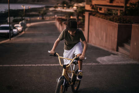 Trendy boy riding his bicycle on the warm light of a summer evening. Young man pedaling with BMX alone on the road near the sea. Teen that fatigue uphill on the bike. Youth recreation positive conceptの写真素材