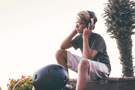 Trendy boy with headphones relaxing listening music after a ride with bicycle. Young male enjoy free time outdoor on a summer evening. Warm filter, exotic background. Youth, recreation, fun, conceptの写真素材