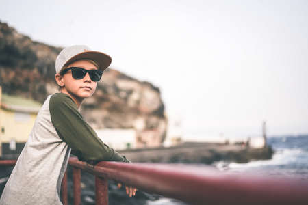 Beautiful smiling teen relaxing leaning against a railing near the sea, looking away with sun glasses and trendy hat. Young boy enjoying last summer days in front of the ocean. Youth, positive conceptの写真素材