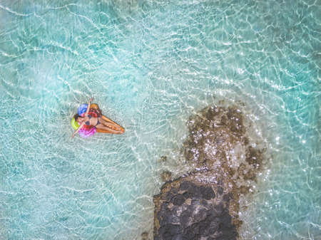 Beautiful girl lying on an ice cream cone mat relaxing during exotic vacation. Woman sunbathing on an inflatable mattress floating on blue tropical water. Zenithal view from drone. Holiday conceptの写真素材