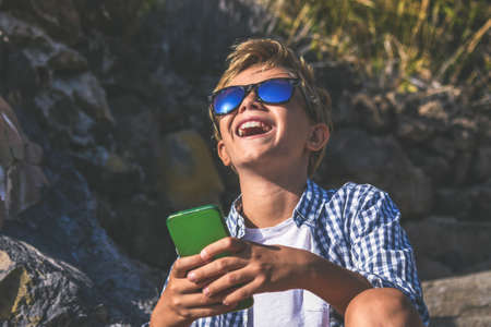 Trendy boy laughing watching social video on smartphone. Happy guy with cheerful expression while texting his friends via social networks using mobile phone, sitting outdoors in the park.の写真素材