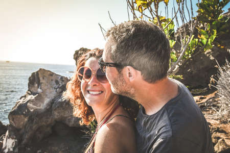 Happy trendy couple having tender moments outdoor. Young lovers having fun in summer vacation smiling and watching the ocean together. Rocks and sea in the background. Travel, love and holiday conceptの写真素材