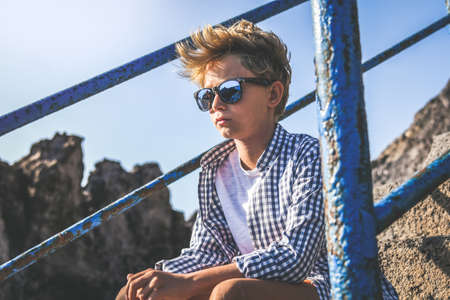 Serious trendy boy sitting on a staircase looking at the horizon waiting friends. Portrait of a thoughtful young male relaxing outdoor. Pensive teen casual clothed enjoying summer holiday alone.の写真素材