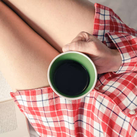 Top view of a female hand holding a cup of tea legs and a book in background. Girl have breakfast sitting on bed with trendy pajama. Square image of a woman reading a book and drinking a mug of coffeeの写真素材