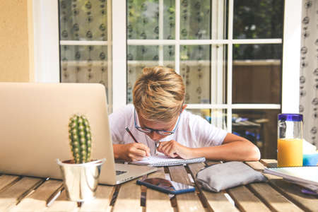 Beautiful young boy doing homework outdoor. Child working with pencil and laptop at home after school. Kid studying on the terrace during holiday drinking orange juice. Caucasian male working with pcの写真素材