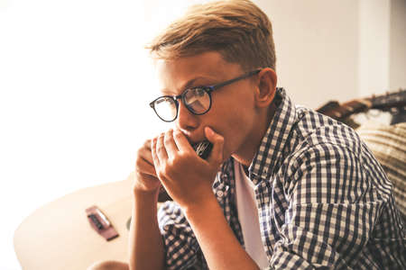Close up portrait of beautiful young boy playing mouth harmonica. Music student exercises alone at home. Trendy caucasian child plays instruments sitting on the sofa. Blonde guy doing musical homeworkの写真素材