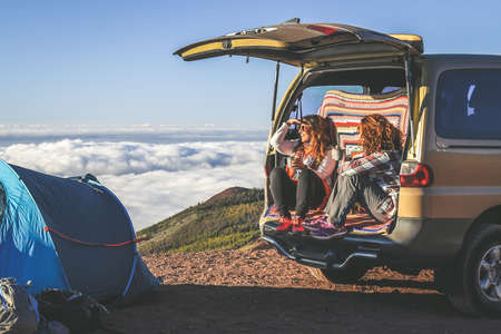 Couple of woman enjoying outdoor sitting on the back of a 4x4 van. Gorgeous mountain panorama on the...