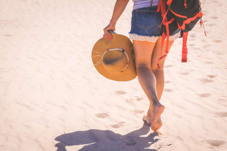 Close up view of a trendy girl walking barefoot on white sand. Jeans shorts and straw hat in her...