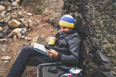 Bearded middle-aged man with woolen hood resting sitting after a trekking day Male drinking coffee looking on cell phone. Relaxing with hot beverage in the National Park. Positive and healthy conceptの写真素材