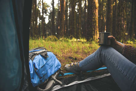 Close up view of hand and legs of a young woman sitting in a tent drinking cup of coffee Trendy girl relaxing in the forest after a trekking holiday Tourism adventure active lifestyle healthy conceptの写真素材