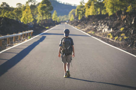 Back view of a young boy walks alone in the middle of the street. Teen with backpack leaving after the end of school Man walking to a new travel adventure. Positive, brave, and adventurous concept.の写真素材