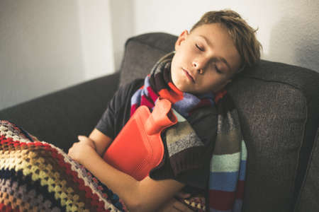 Sick boy sitting with woolen blanket and hot water bottle. Sad teen with the flu rests alone at home...