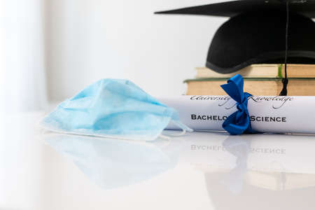 Closeup of graduation hat with degree and surgical mask on white background. On line virtual celebration at home for students during pandemic coronavirus. Graduation in lockdown. Graduating concept.の写真素材